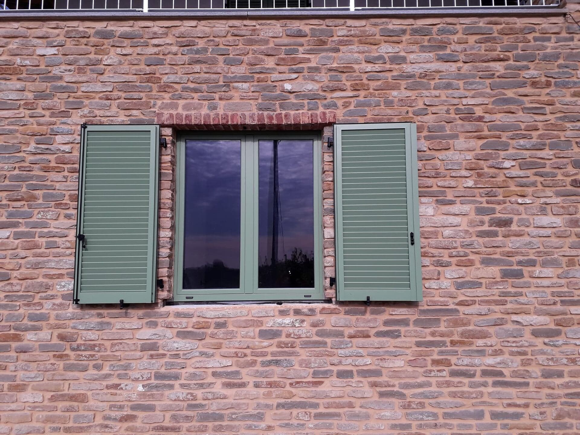 Finestra con le persiane verde salvia aperte su un muro in mattoni a vista, con il cielo riflesso nei vetri