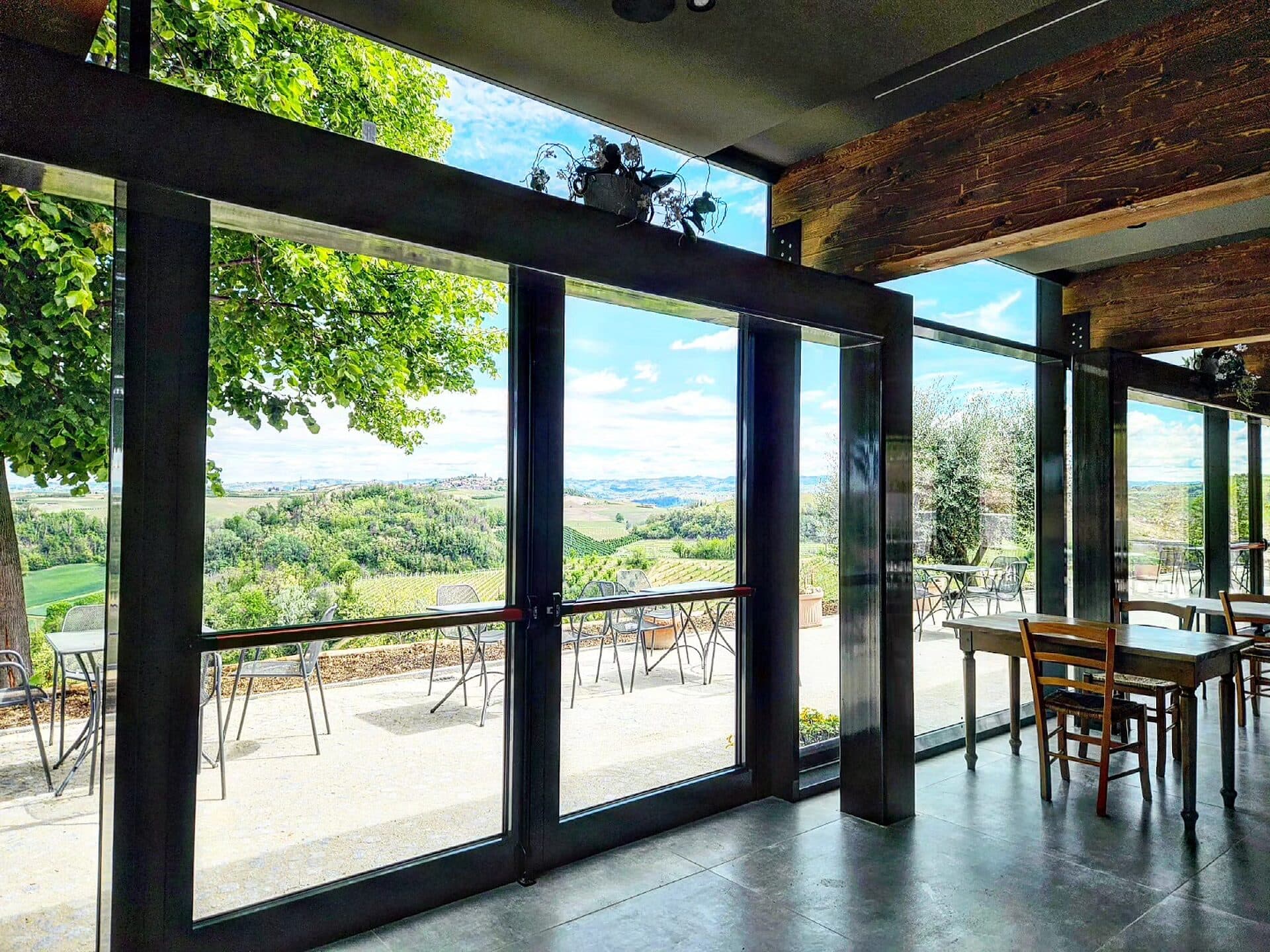 Veranda con pareti vetrate scorrevoli in alluminio scuro sotto una copertura con travi in legno, aperta sulle colline coltivate a vigneto
