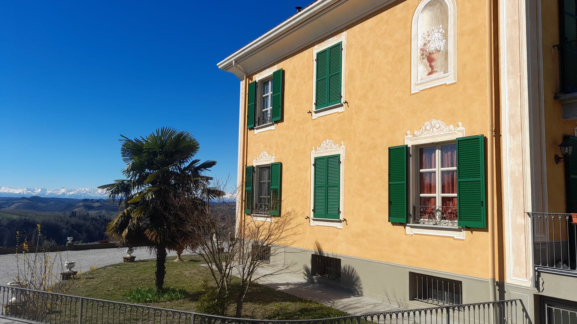 La villa gialla con le persiane verdi vista dal prato, con la palma davanti e le Alpi innevate all'orizzonte