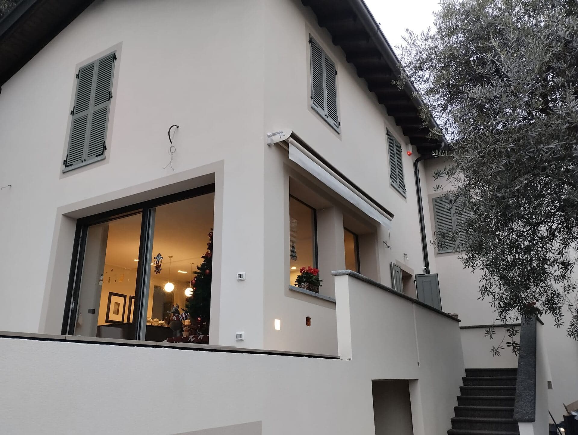 La villa all'imbrunire con le finestre illuminate, le persiane verde salvia accostate e l'olivo sul fianco